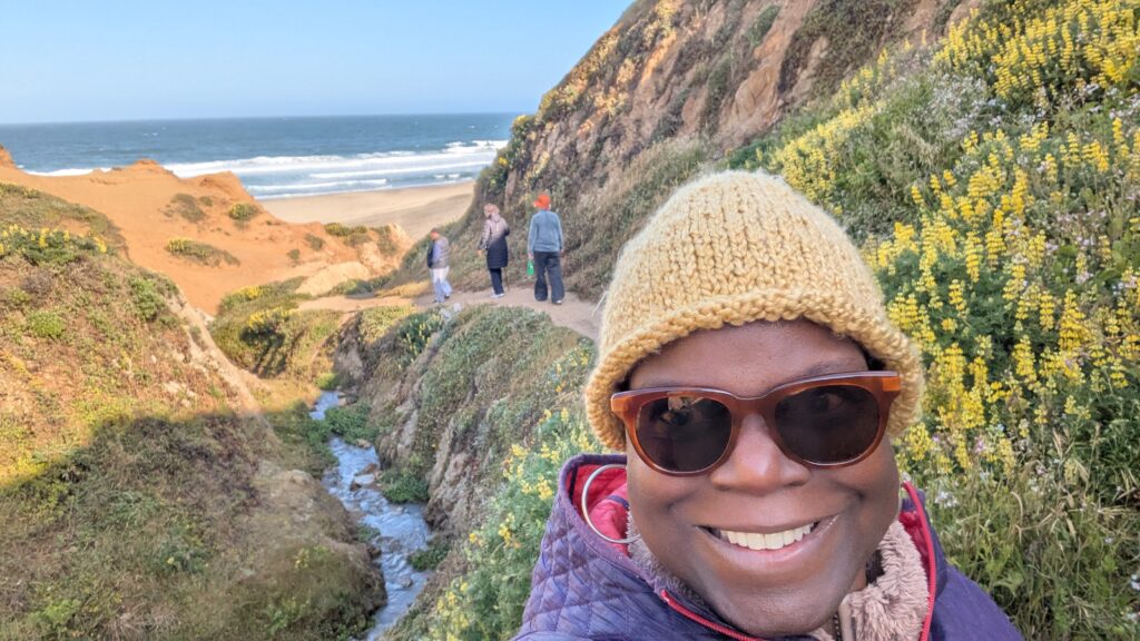 Dr. Abigail bundled in hat and warm coat with hills of flowers and the ocean peaking through inthe background
