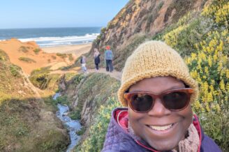 Dr. Abigail bundled in hat and warm coat with hills of flowers and the ocean peaking through inthe background
