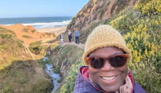 Dr. Abigail bundled in hat and warm coat with hills of flowers and the ocean peaking through inthe background